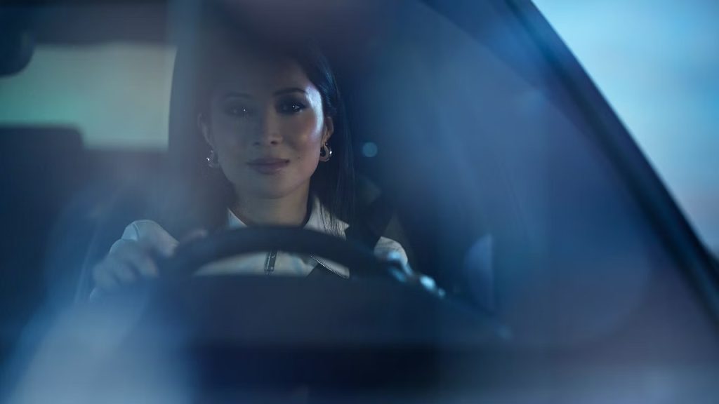 Photo of a woman in the driver’s seat, seen through the front windshield with hands on the wheel.
